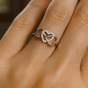 May include: A silver ring featuring two intertwined heart shapes. One heart is encrusted with small, sparkling clear stones. The ring is shown on a person's finger, with a soft background.