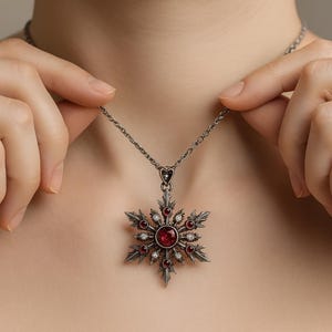 Könnte beinhalten: Eine silberfarbene Schneeflocken-Anhänger-Halskette mit einem großen, runden, roten Edelstein in der Mitte und kleineren roten und klaren Edelsteinen. Der Anhänger hängt an einer filigranen Kette.