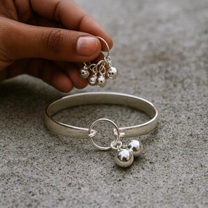 Pode incluir: Um conjunto de pulseira e brincos de prata. A pulseira é uma faixa simples com um pingente circular e duas esferas de prata penduradas. O brinco correspondente tem um aro circular com várias esferas de prata penduradas. As joias estão em uma superfície cinza.
