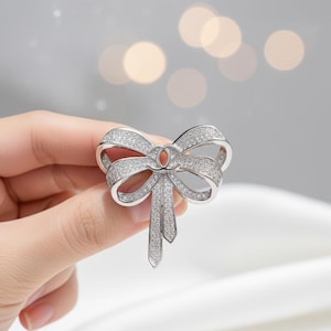 May include: A silver bow brooch, adorned with numerous small, sparkling crystals. The brooch has a detailed bow design with two hanging ribbon-like elements. The brooch is held in a hand against a soft, blurred background.