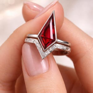 May include: A two-piece ring set featuring a silver band with a V-shaped design and a large, red, geometric gemstone. The band is adorned with small, clear stones and delicate patterns. The gemstone is a vibrant, deep red color.
