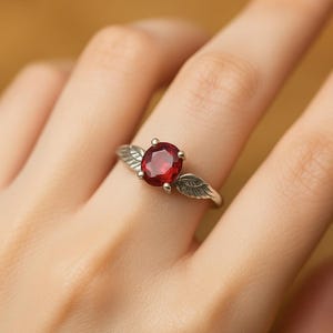 May include: A silver ring with a large, round, red gemstone. The ring's band has a wing design on either side of the gemstone. The ring is displayed on a finger.