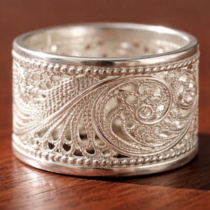 Op de afbeelding: Een zilveren filigraan ring met een ingewikkeld open ontwerp. De ring heeft een gedetailleerd patroon van wervelende lijnen en delicate uitsparingen, waardoor een kantachtig uiterlijk ontstaat. De band is ongeveer 2.5 cm breed.