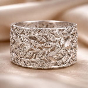 Può includere: Un anello largo color argento con un intricato motivo a foglie. L'anello è ricoperto di piccole pietre scintillanti e le foglie sono disposte in fasce orizzontali. L'anello poggia su una superficie beige chiaro.