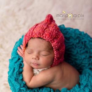 Può includere: Un neonato con un berretto di lana rosa dorme serenamente in una coperta turchese con volant. Il bambino è rannicchiato nella coperta, con le braccia sotto il mento. L'immagine è dolce e serena, catturando l'innocenza di un neonato.
