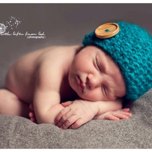 Puede incluir: Un bebé recién nacido durmiendo con un gorro de punto turquesa con un botón de madera en la parte superior. El bebé está acostado sobre una manta gris.