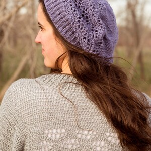 May include: A woman wearing a purple crocheted beanie with a lacy design. She is wearing a grey crocheted sweater with a lacy design on the back. The sweater has the text "www.melodays-makings.com" on the bottom.