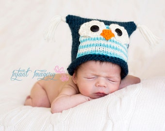 May include: A newborn baby wearing a blue and white knitted owl hat with black button eyes and an orange beak. The baby is lying on a white blanket.
