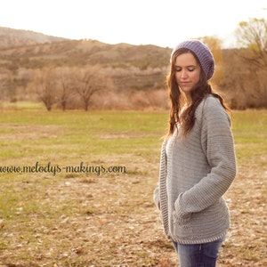 May include: A woman wearing a light grey crocheted cardigan with a purple beanie. The cardigan has a deep V-neck and long sleeves. The woman is standing in a field with a blurred background.