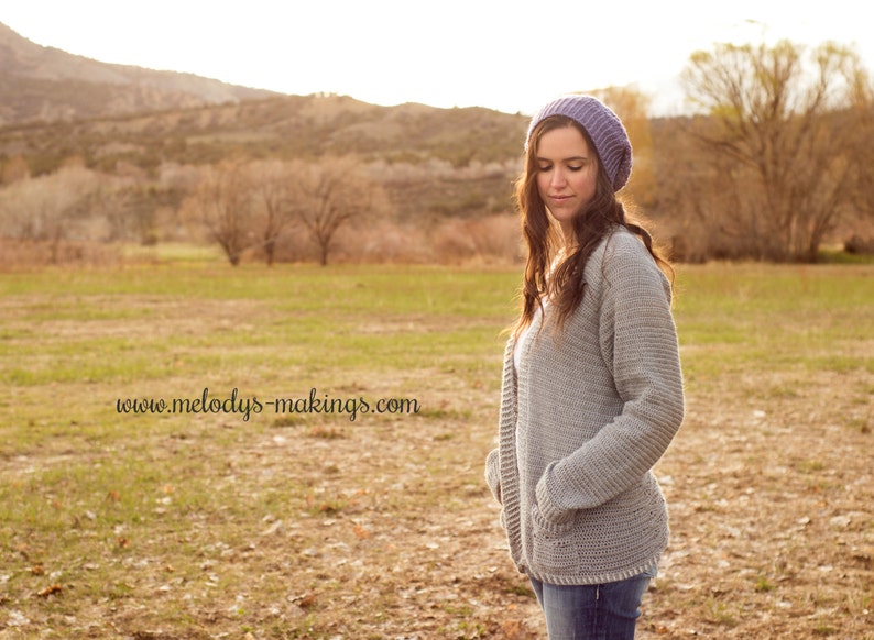 May include: A woman wearing a light grey crocheted cardigan with a purple beanie. The cardigan has a deep V-neck and long sleeves. The woman is standing in a field with a blurred background.