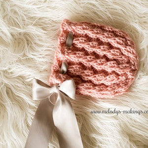May include: A pink knitted baby bonnet with a cable knit pattern. The bonnet features two grey ribbon accents and a large white satin bow. It rests on a white fluffy surface.