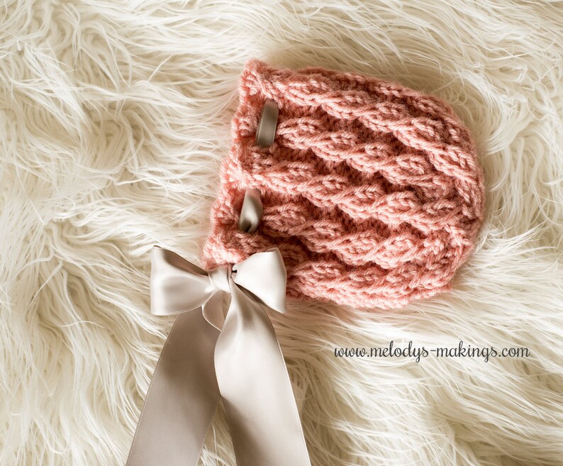 May include: A pink knitted baby bonnet with a cable knit pattern. The bonnet features two grey ribbon accents and a large white satin bow. It rests on a white fluffy surface.