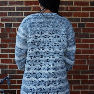 May include: A long-sleeved, crocheted sweater with a white and grey colour pattern. The sweater has a loose fit and a textured design.