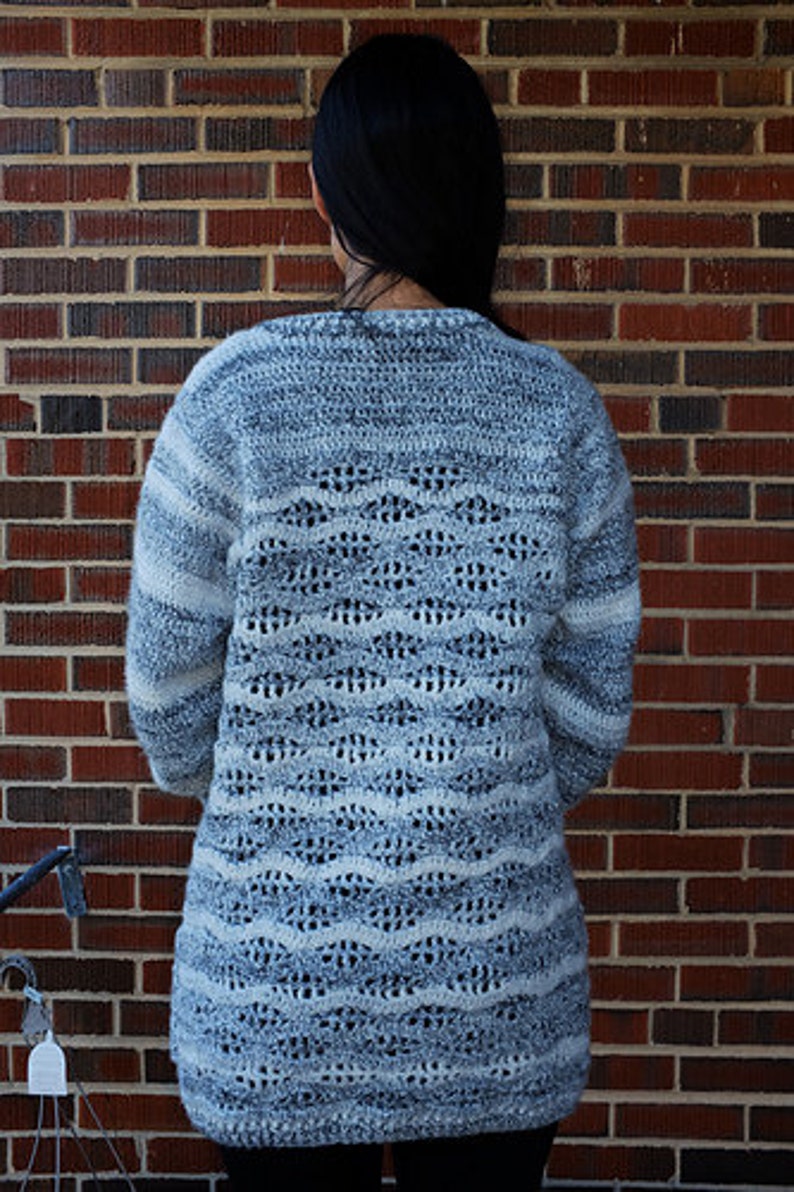 May include: A long-sleeved, crocheted sweater with a white and grey colour pattern. The sweater has a loose fit and a textured design.