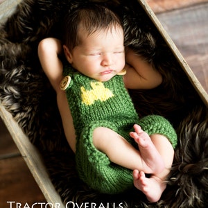 May include: A newborn baby wearing a green knitted tractor overalls outfit with yellow and brown details. The baby is sleeping in a wooden crate lined with brown fur.
