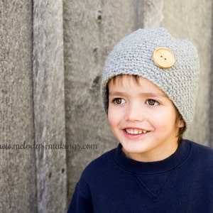 Puede incluir: Un gorro gris tejido a ganchillo con un botón de madera grande. El gorro lo lleva una persona que lleva una sudadera azul marino. La dirección del sitio web www.melodys-makings.com es visible en la imagen.
