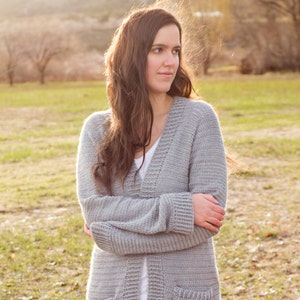 May include: A grey crocheted cardigan with a V-neck and pockets. The cardigan is being worn by a person with long brown hair, standing in a field.