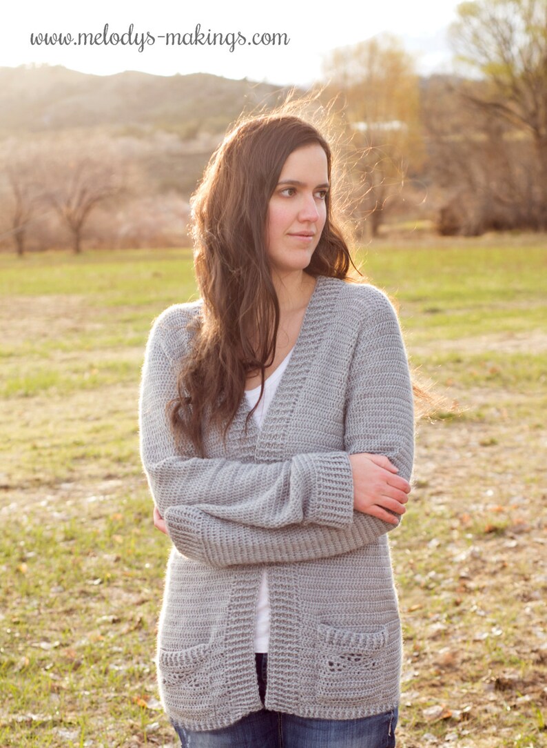 May include: A grey crocheted cardigan with a V-neck and pockets. The cardigan is being worn by a person with long brown hair, standing in a field.