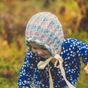 May include: A child wearing a knitted bonnet in shades of blue, pink, and white, with a cream-coloured ribbon tie. The child is wearing a blue top with a white pattern and blue jeans. The image has a soft, natural background.