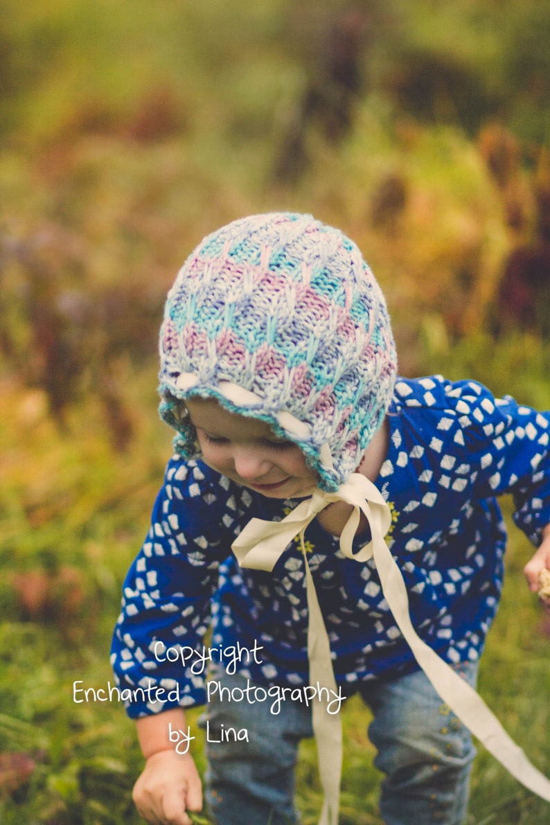 May include: A child wearing a knitted bonnet in shades of blue, pink, and white, with a cream-coloured ribbon tie. The child is wearing a blue top with a white pattern and blue jeans. The image has a soft, natural background.