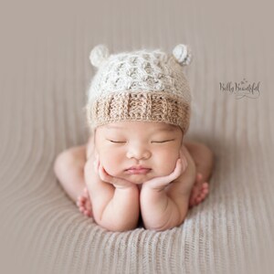 Puede incluir: Un bebé recién nacido con un gorro de punto beige y blanco con orejas de oso, durmiendo con las manos debajo de la barbilla. El bebé está acostado sobre una manta de punto beige.