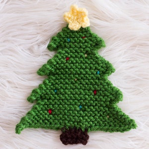 Op de afbeelding: Een groene gebreide kerstboomversiering met een gele ster bovenop en rode en blauwe lampjes. De boom ligt op een wit, pluche oppervlak.