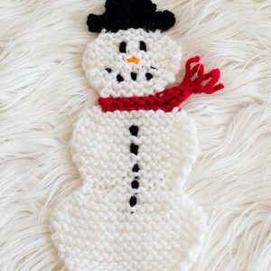 Puede incluir: Decoración de muñeco de nieve hecha a mano. El muñeco de nieve está hecho de hilo blanco, con un sombrero de copa negro, ojos y boca de carbón y una nariz de zanahoria naranja. Una bufanda roja está alrededor del cuello. Botones negros recorren el centro del cuerpo.