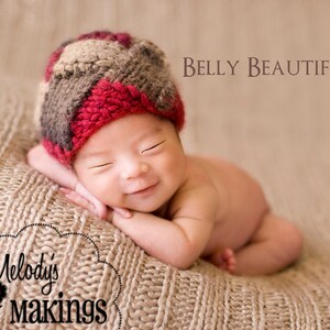 May include: A baby wearing a red, brown, and beige knitted hat with a happy expression. The baby is lying on a beige knitted blanket. The text "Belly Beautiful" is visible in the upper right corner of the image.