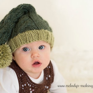 Puede incluir: Un bebé con un gorro de elfo tejido en verde y beige con un pompón a juego. El bebé también lleva un chaleco marrón de ganchillo sobre una camiseta blanca. El bebé tiene ojos azules y mira a la cámara.