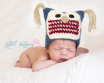 May include: A baby wearing a blue and red owl-shaped knitted hat with white accents. The baby is lying on a white blanket and has their eyes closed.