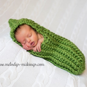 May include: A newborn baby is sleeping soundly in a green crocheted pea pod-shaped swaddle. The baby is wearing a white hat and has a peaceful expression on their face.