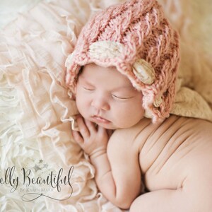 May include: A sleeping baby wearing a pink cable-knit hat with cream-coloured accents. The hat has a flower-like embellishment. The baby is lying on a soft, ruffled, cream-coloured blanket. The image has a soft, warm colour palette.