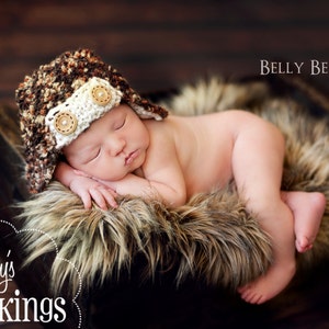 May include: A newborn baby wearing a brown and white crocheted aviator hat with brown buttons, sleeping on a brown faux fur rug in a wooden crate. The text "Belly Beautiful" is visible in the upper right corner of the image.