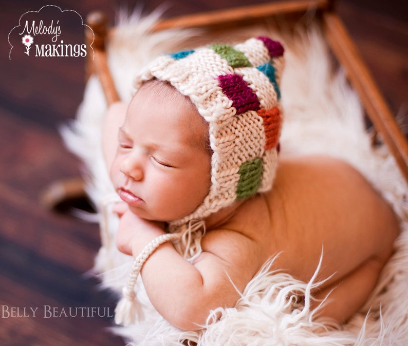 May include: A newborn baby wearing a white, green, orange, and purple crocheted hat with a white fur background. The baby is sleeping with their eyes closed and their arms tucked under their chin.