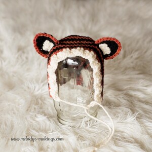 May include: A crocheted tiger hat with black, white, and orange stripes. The hat has two floppy ears and a white crocheted brim.