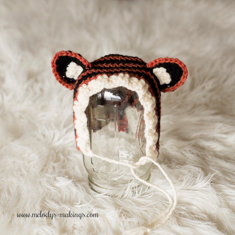 May include: A crocheted tiger hat with black, white, and orange stripes. The hat has two floppy ears and a white crocheted brim.