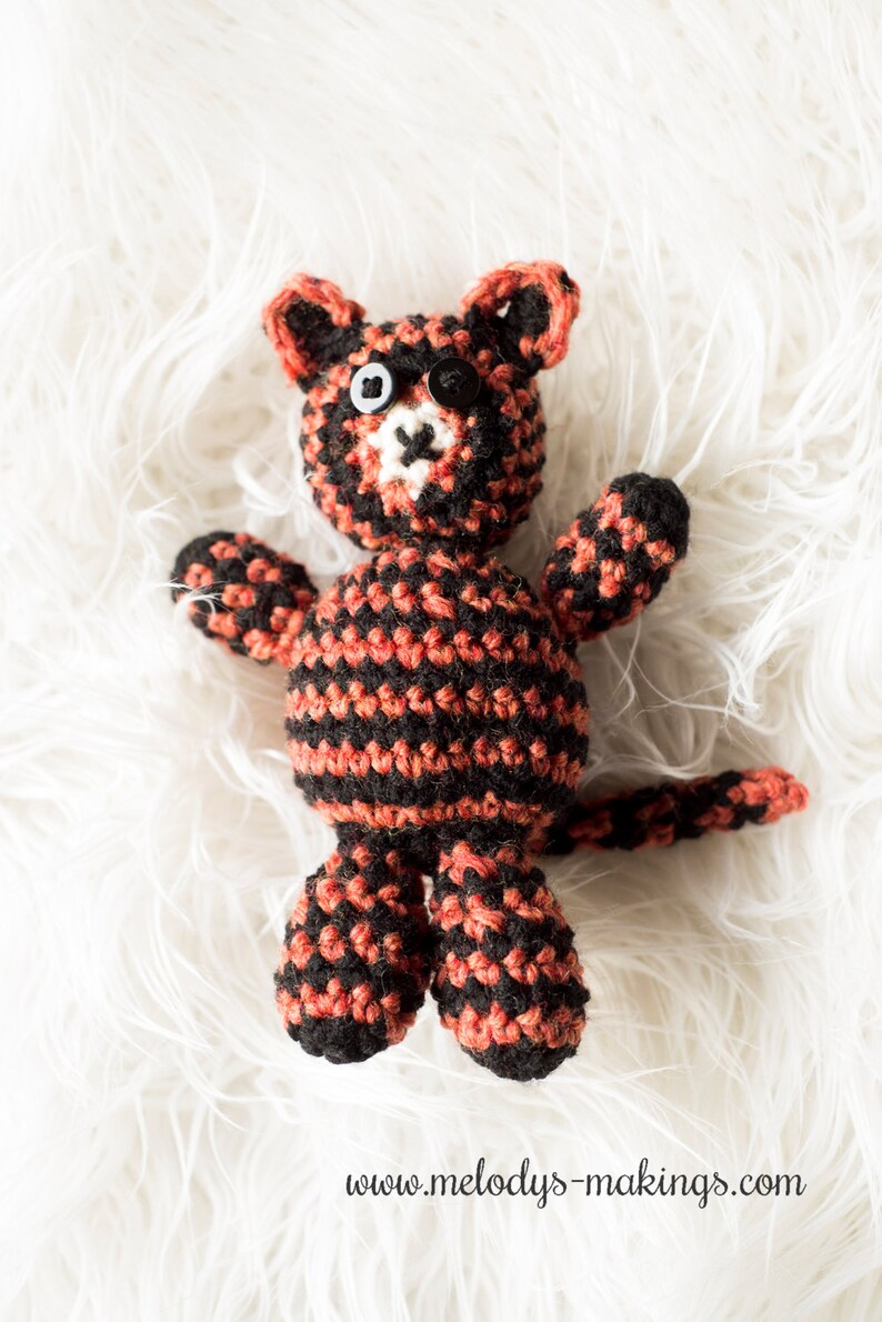 May include: A crocheted cat toy with black and orange stripes. The cat has black button eyes and a white nose.