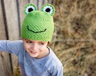 May include: A green crocheted frog hat with black eyes and a black smile. The hat is being worn by a child wearing a grey shirt.