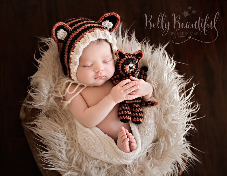 May include: A newborn baby wearing a brown and white striped crocheted hat with ears, sleeping in a white fur-lined basket. The baby is holding a brown and black crocheted animal toy.