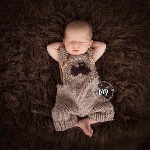 May include: A newborn baby wearing a brown knitted outfit with a tractor design on the front, lying on a brown fuzzy blanket.