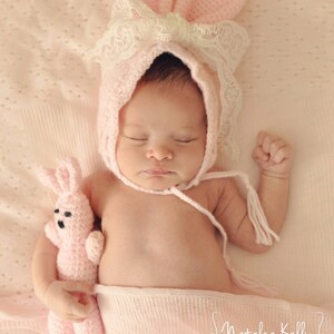 May include: A sleeping baby wearing a pink crocheted bunny hat with white lace trim and holding a pink crocheted bunny toy. The baby is lying on a pink blanket with a white polka dot pattern.
