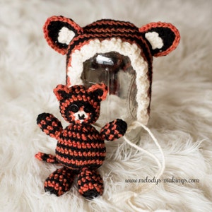 May include: A crocheted tiger hat with black and orange stripes and white trim. A small crocheted tiger toy with black and orange stripes is sitting in front of the hat. The hat and toy are on a white fluffy surface. www.melody's-makings.com