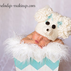 May include: A newborn baby wearing a white crocheted lamb hat with blue accents and black button eyes. The baby is lying in a white fluffy basket with a blue and white chevron pattern.