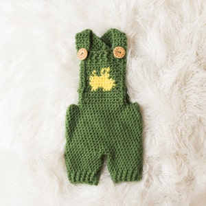 May include: Green crocheted baby overalls with yellow bunny applique and wooden buttons.