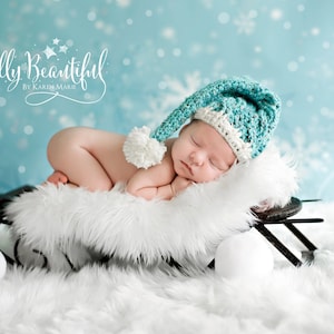 May include: A newborn baby wearing a teal and white crocheted hat with a pom-pom, sleeping on a white fur-covered sled. The background is a light blue with snowflakes and the text "Belly Beautiful by Karen Marie".