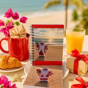 May include: A spiral-bound notebook with a red cover and a Santa Claus figurine on the beach. A red mug with a gift box, a croissant, and a glass of orange juice are also present. The scene is set on a white surface with pink flowers and gold stars.