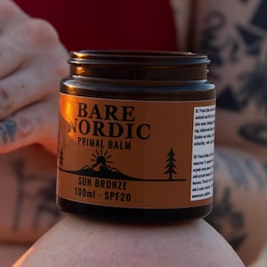 Könnte beinhalten: Ein dunkelbraunes Glastiegelchen mit Bare Nordic Primal Balm Sun Bronze SPF20. Das Etikett zeigt eine Berg- und Baumgrafik mit dem Text Bare Nordic, Primal Balm, Sun Bronze und 100ml - SPF20. Der Tiegel hat einen schwarzen Schraubdeckel.