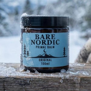 Puede incluir: Un tarro de cristal de Bare Nordic Primal Balm, con tapa negra y etiqueta azul claro. La etiqueta presenta el texto "Bare Nordic Primal Balm", "Original" y "100ml", junto con un gráfico de montaña y árbol. El tarro está rodeado de hielo.