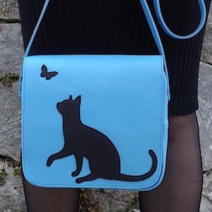 May include: A light blue crossbody bag with a black cat silhouette and a butterfly. The bag has a matching blue strap and is worn over the shoulder. The cat is reaching up towards the butterfly.