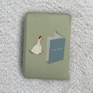 Peut inclure: Un étui rectangulaire vert sauge avec un dessin d'oie portant un bonnet de Père Noël, lisant un livre avec le texte "not smut". L'oie est blanche avec un bec jaune et des accents rouges.